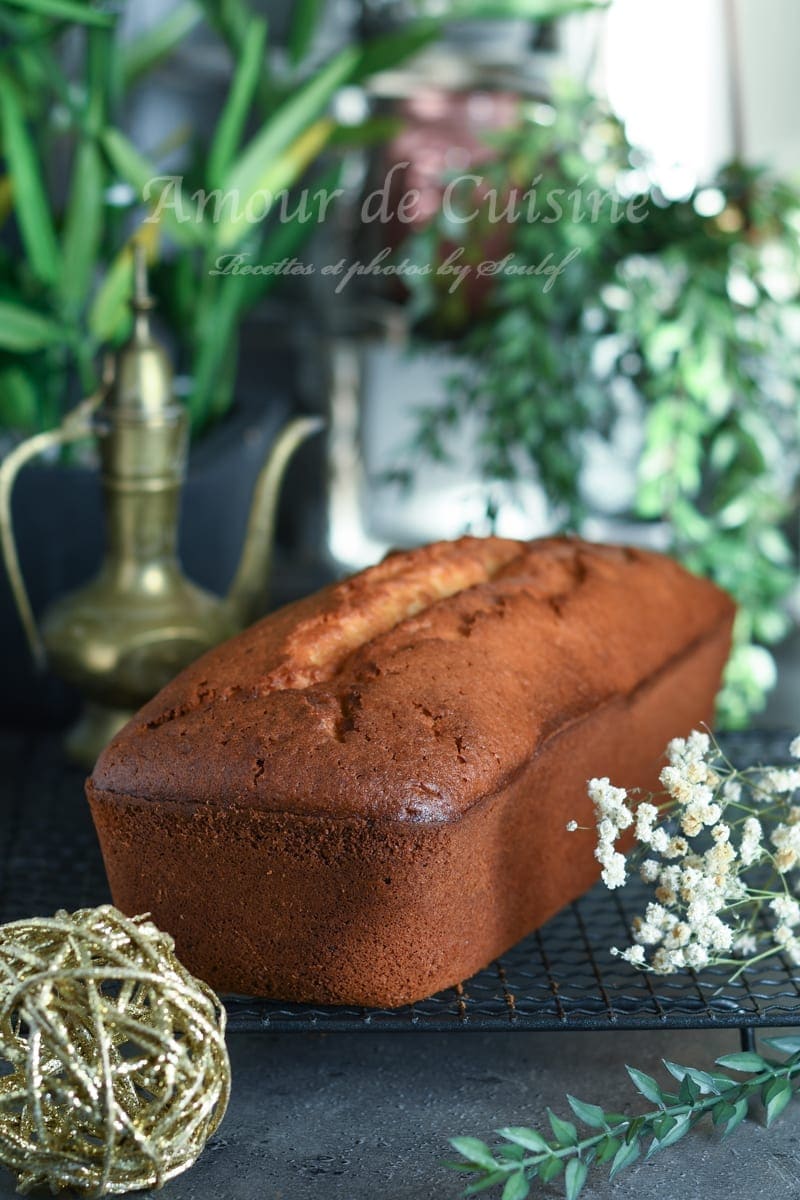 Golden-brown yogurt loaf cake cooling on a wire rack, photographed whole with a cracked top, surrounded by greenery and small white flowers in a softly blurred background.