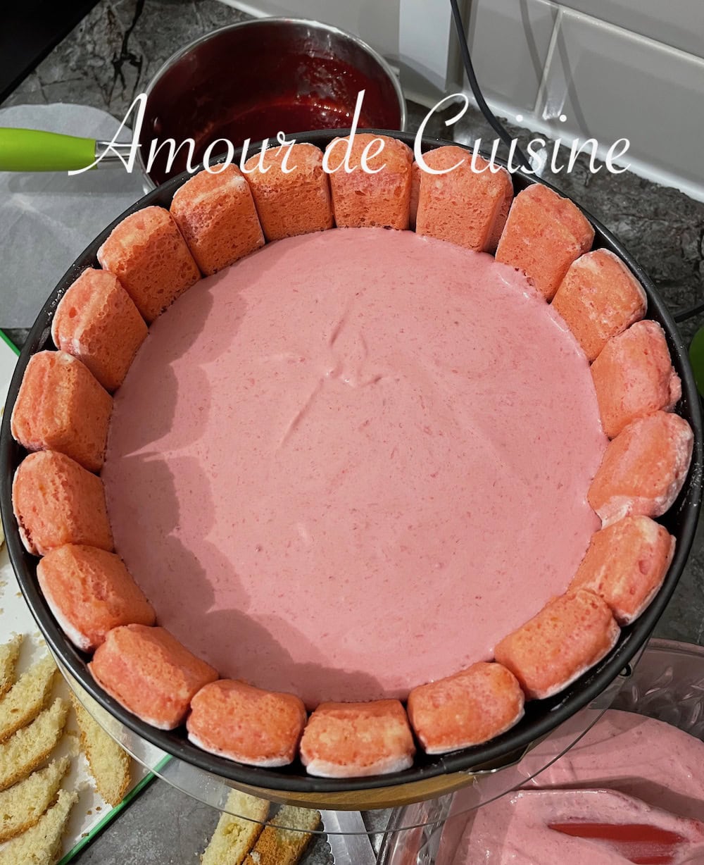 Charlotte aux fraises en cours de préparation, garnie de mousse rose aux fraises et entourée de biscuits roses de Reims, disposés debout dans un moule à charnière sur un plan de travail avec des ustensiles de cuisine visibles en arrière-plan