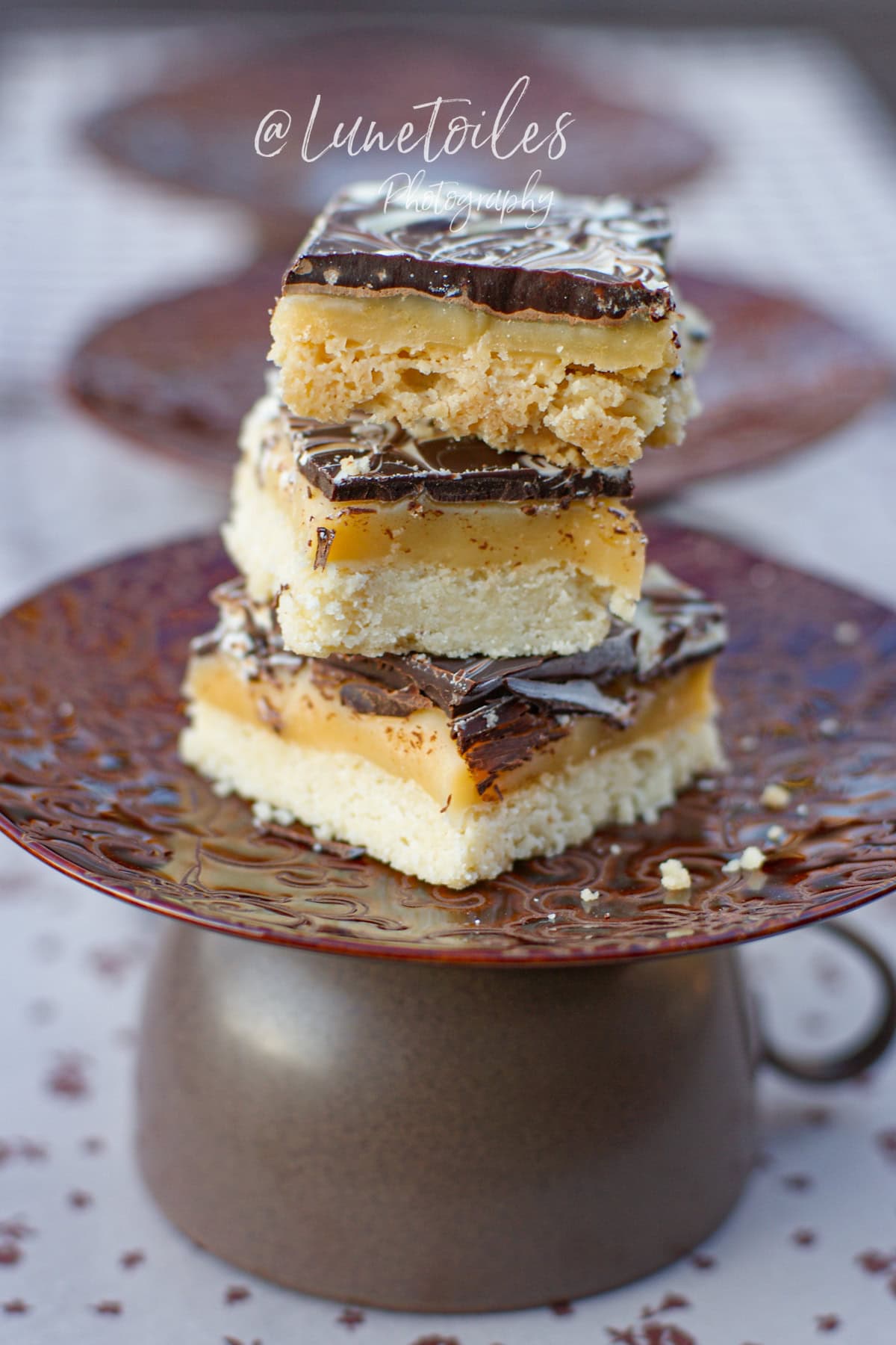 Empilement de trois carrés de shortbread millionnaire avec base sablée, caramel fondant et chocolat marbré, présenté sur une assiette vintage surélevée
