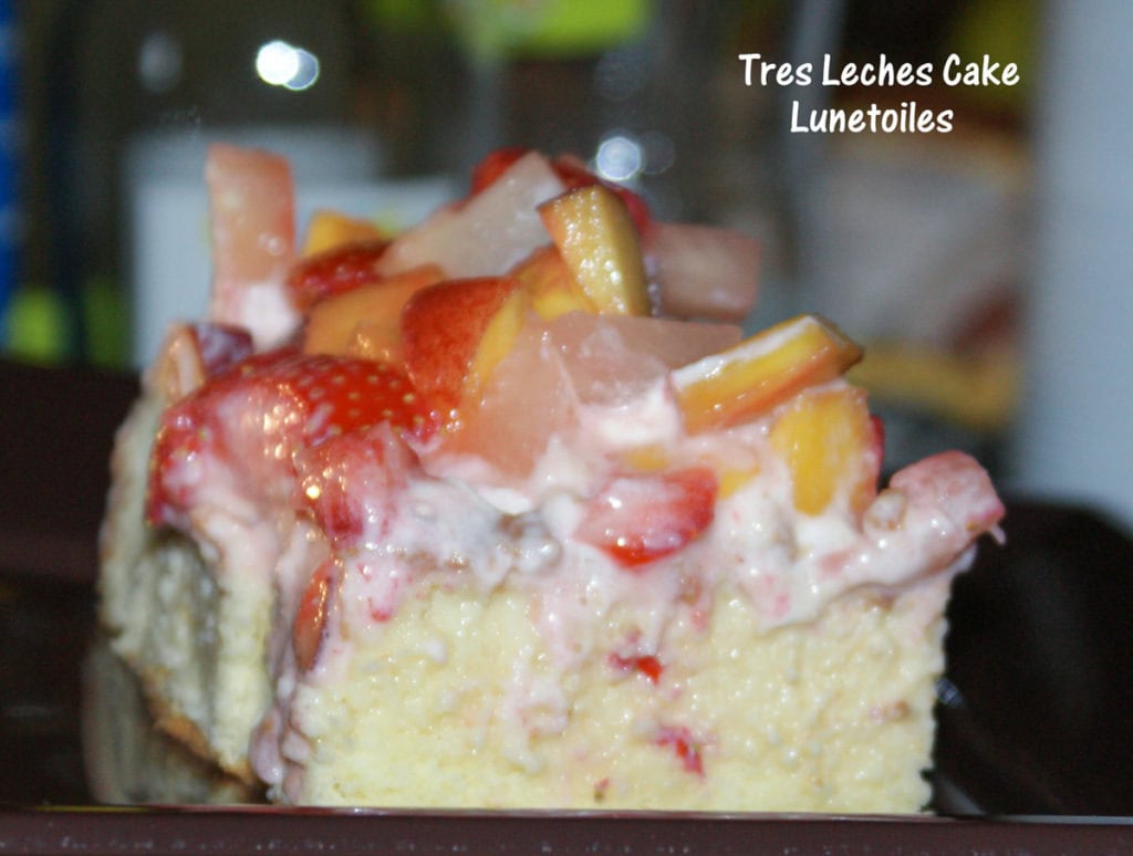 Close-up of a slice of Tres Leches cake topped with whipped cream and diced fresh fruits (strawberries, mango, pear), served on a brown plate.