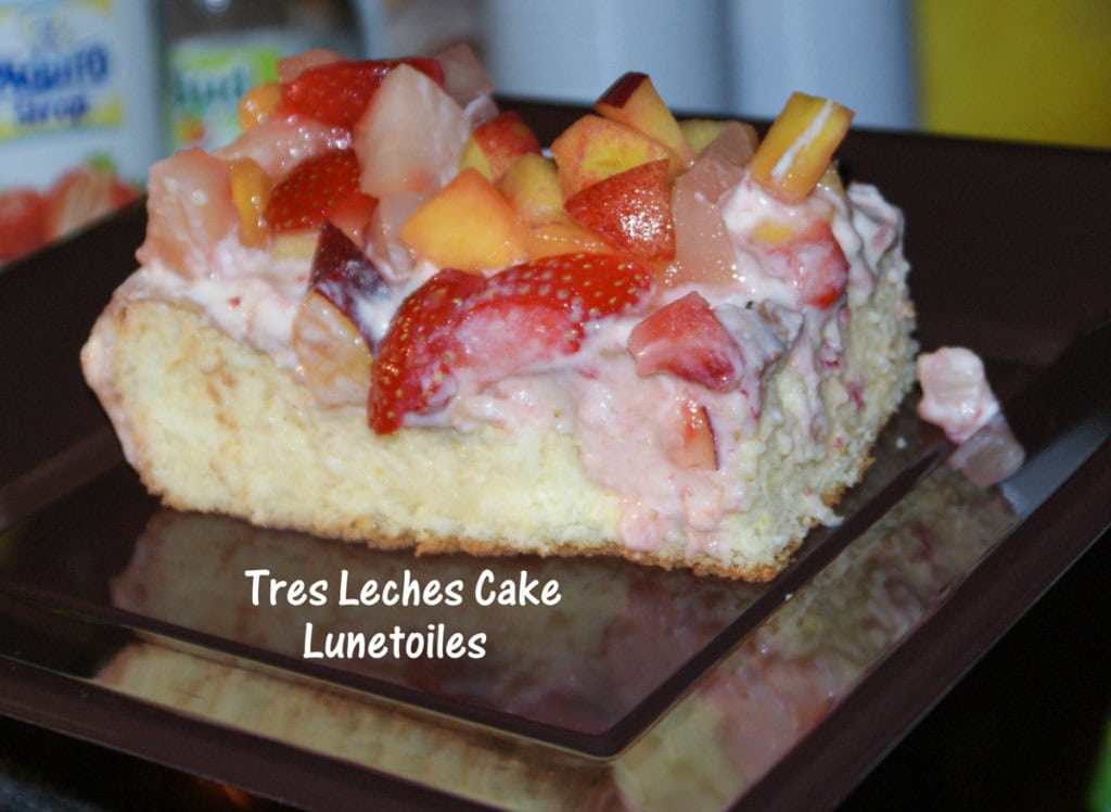 Close-up of a slice of soaked Tres Leches cake topped with whipped cream and assorted fresh fruit pieces (strawberries, mango, pear), served on a brown plate.