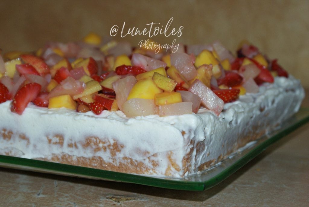 Rectangular Tres Leches cake topped with whipped cream and generously garnished with diced fresh fruits (strawberries, mango, pear), served on a glass platter.