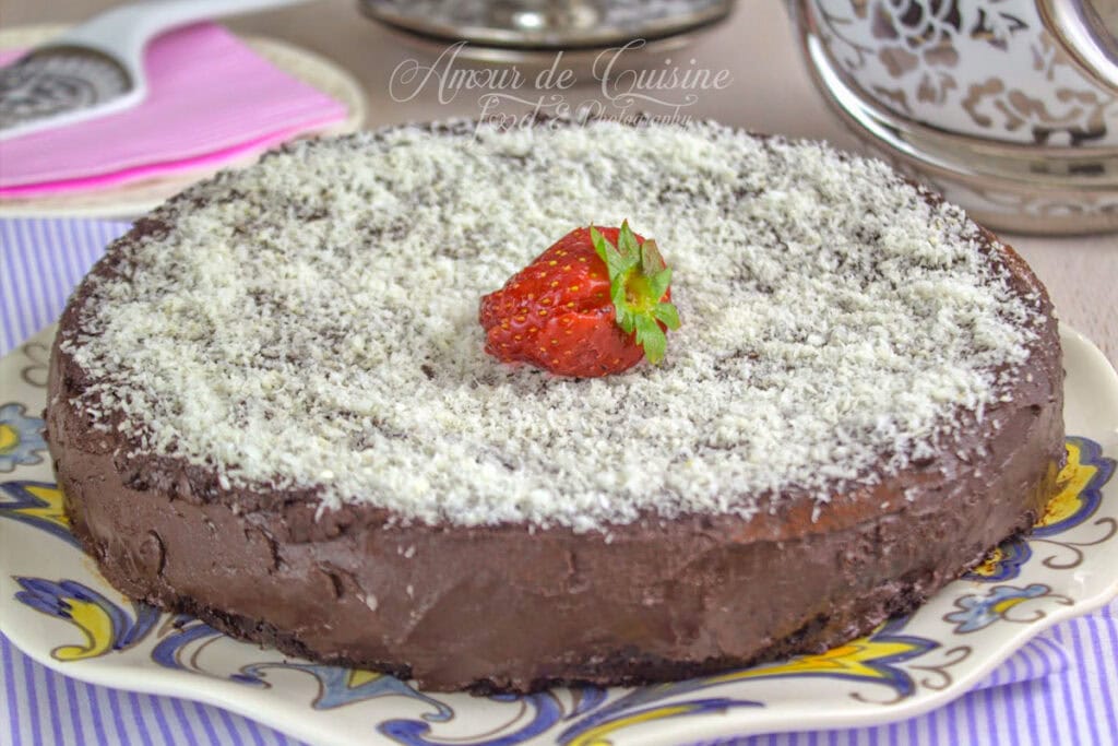 gâteau au yaourt au chocolat facile recouvert de ganache au chocolat, saupoudré de noix de coco râpée et décoré d’une fraise, présenté sur un plat à motifs.