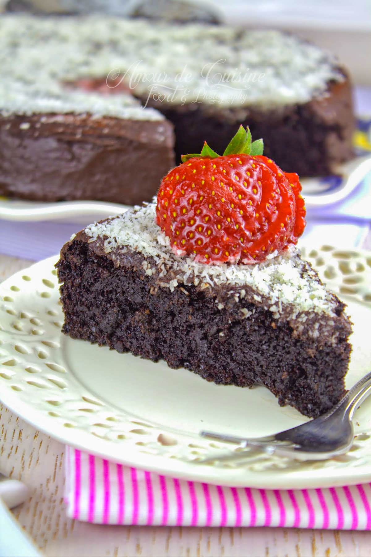 part de gâteau au yaourt au chocolat moelleux nappé de ganache, saupoudré de noix de coco râpée et décoré de fraises fraîches, servie sur une assiette blanche ajourée.