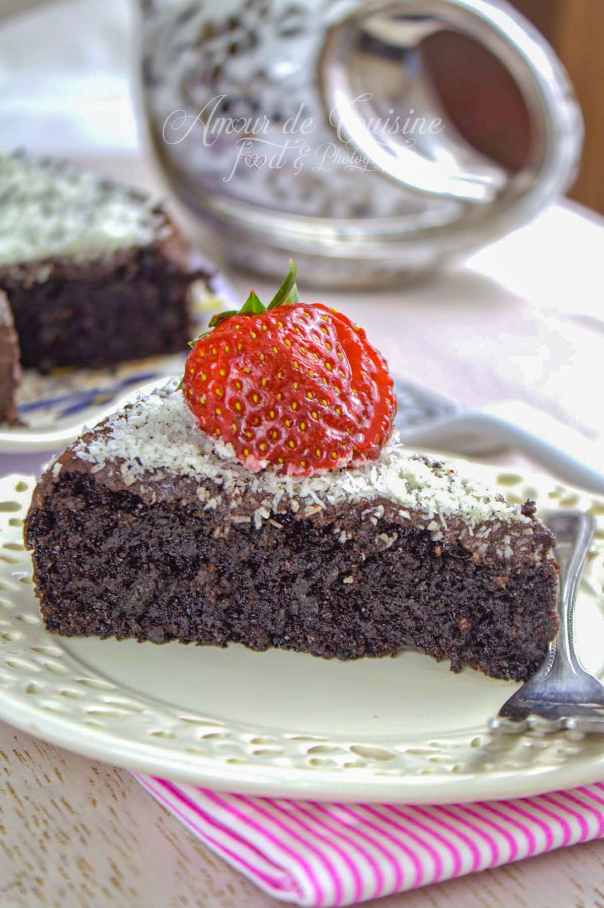 part de gâteau au yaourt au chocolat fondant recouverte de ganache et de noix de coco râpée, garnie d’une fraise fraîche, servie sur une assiette blanche avec une fourchette.