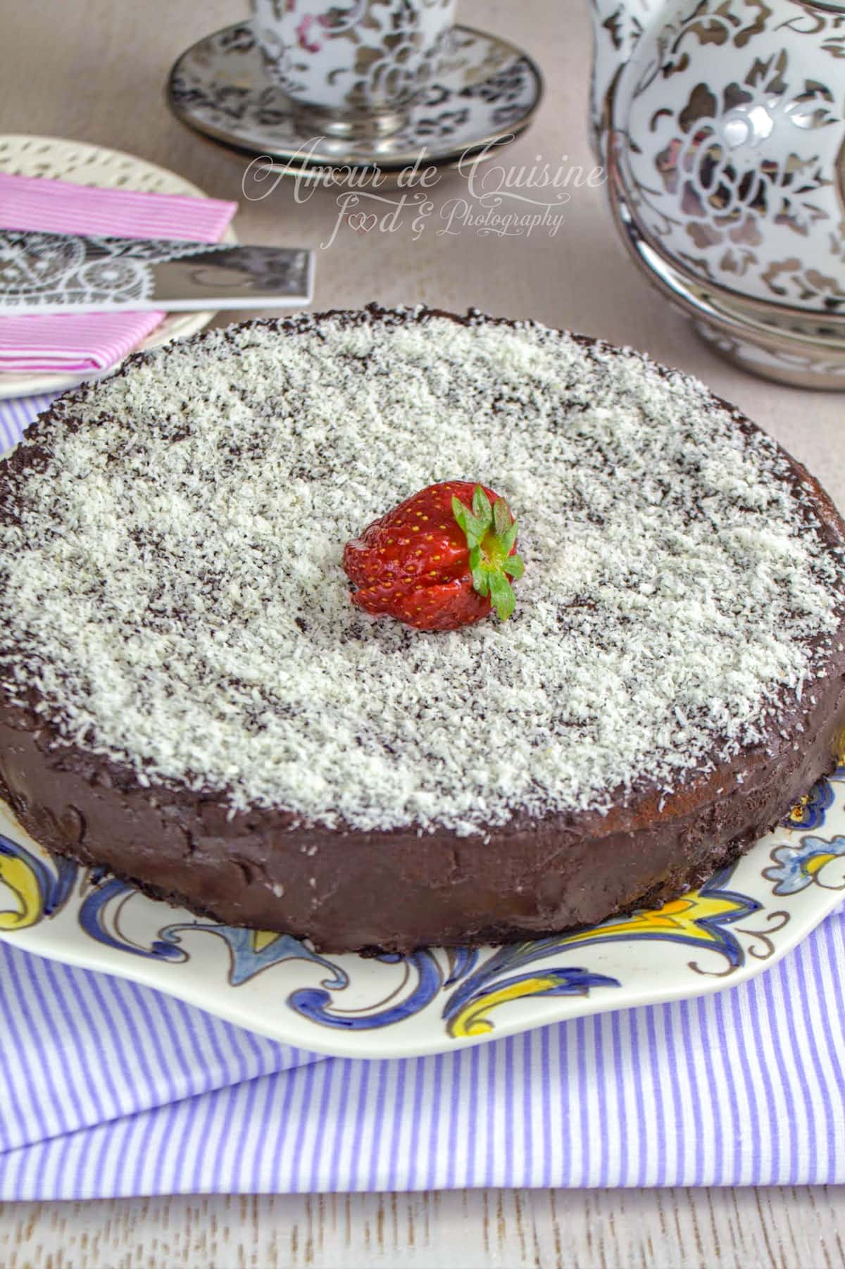 gâteau au yaourt au chocolat recouvert de ganache au chocolat, saupoudré de noix de coco râpée et décoré d’une fraise au centre, présenté sur un plat à motifs.