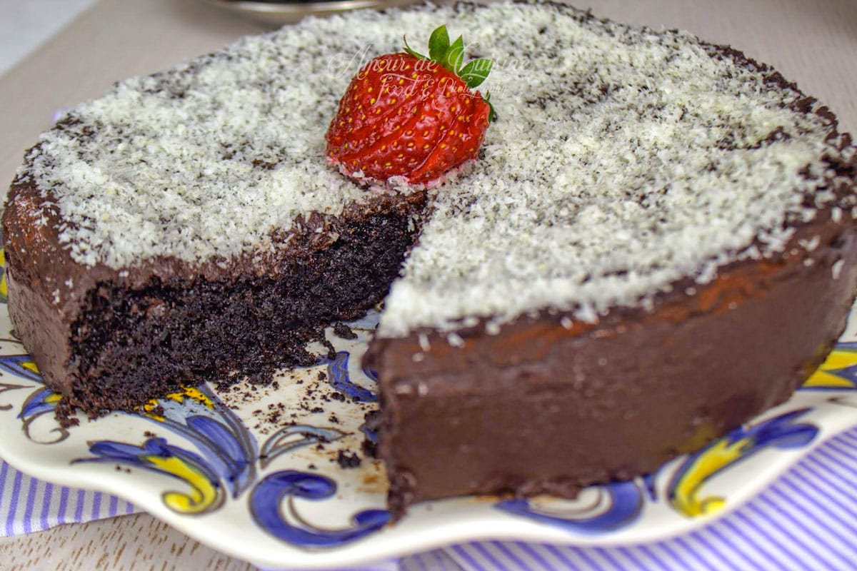 gâteau au yaourt au chocolat moelleux recouvert de ganache et de noix de coco râpée, présenté coupé avec une part retirée, décoré d’une fraise au centre.
