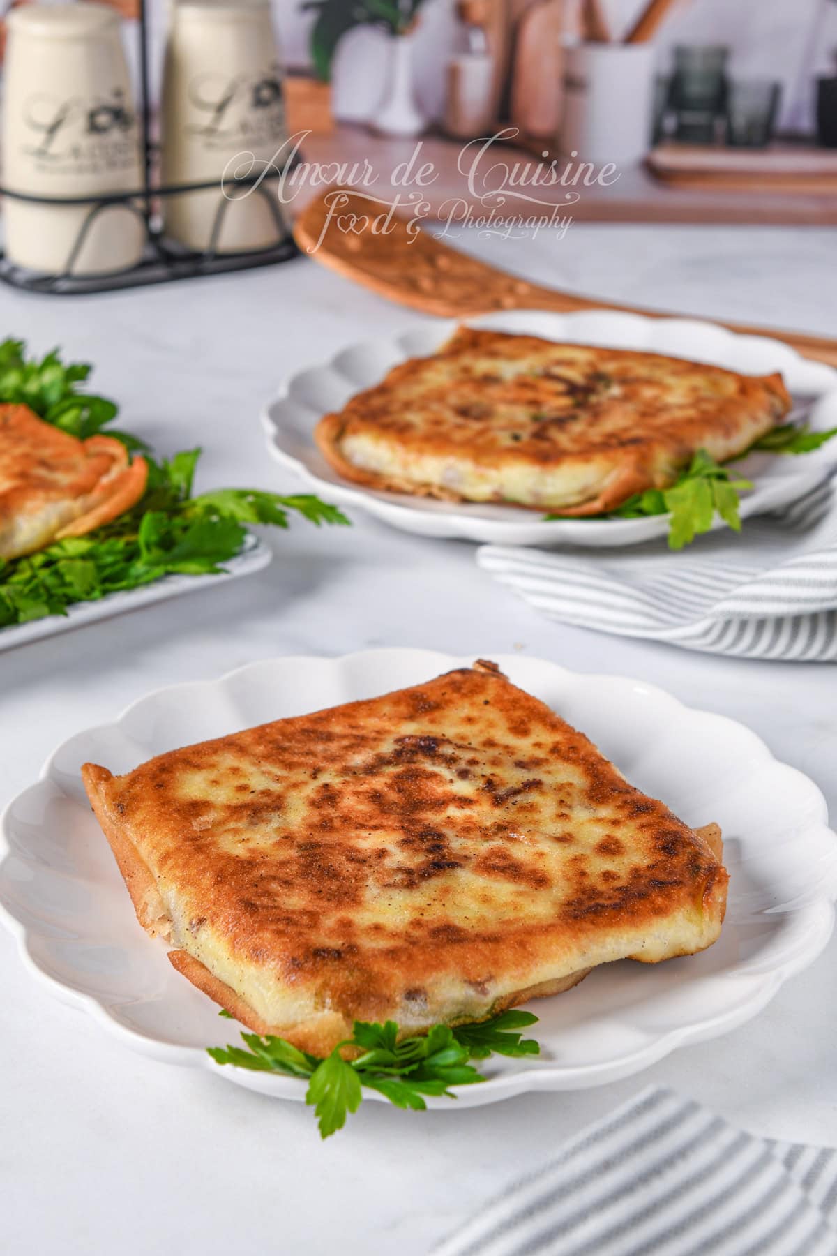 Golden Annabi bourek square on a white plate, with more squares in the background, served with a few parsley leaves on a bright tabletop.