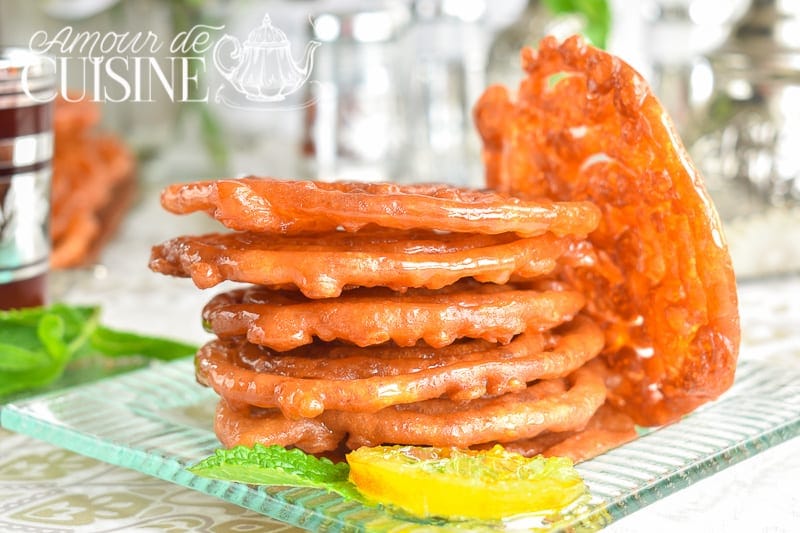 Stack of golden zlabia glazed with honey syrup on a glass platter, served with an orange slice and mint leaves, with one zlabia standing in the background.
