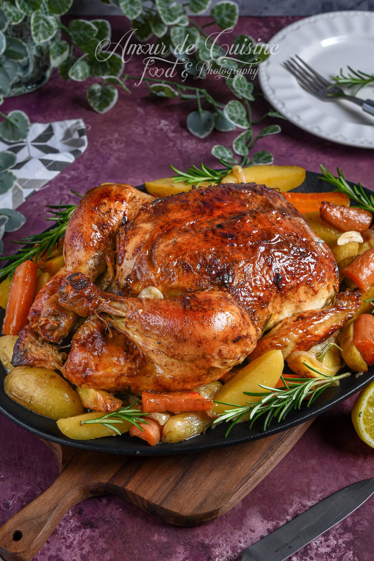 Whole roast chicken with golden, crispy skin, served with roasted potatoes and carrots, garnished with fresh rosemary sprigs on a cast iron dish.