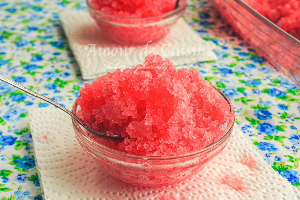Close-up of a glass bowl filled with bright red grenadine granita, served with a spoon on a floral tablecloth, refreshing homemade frozen dessert made with pomegranate syrup.