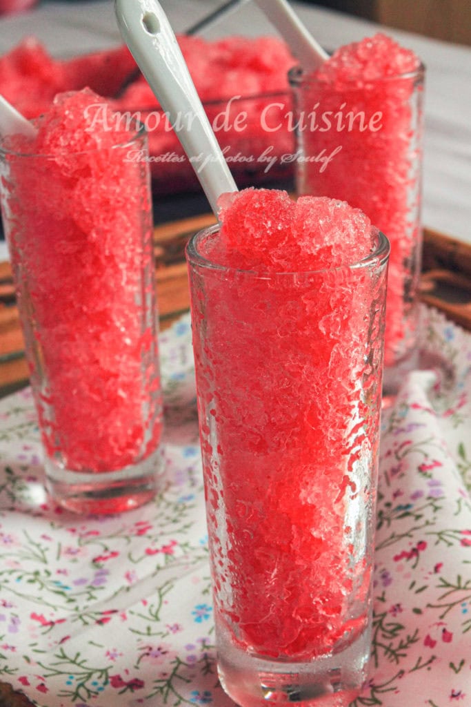 Tall glasses filled with bright red grenadine granita topped with ceramic spoons, placed on a floral-patterned cloth, refreshing homemade frozen dessert made with pomegranate syrup