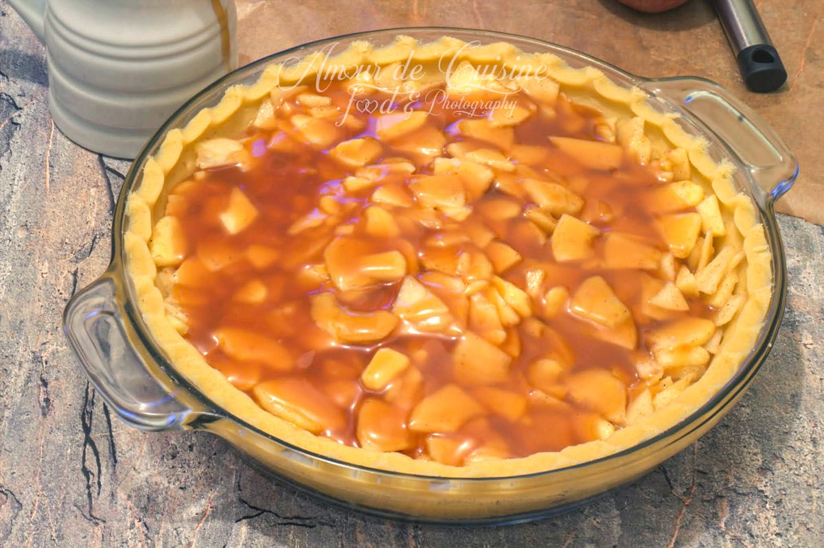 Apple pie d’automne au caramel au beurre salé : fond de pâte sablée garni de pommes émincées nappées de caramel, tarte avant cuisson dans un plat en verre.