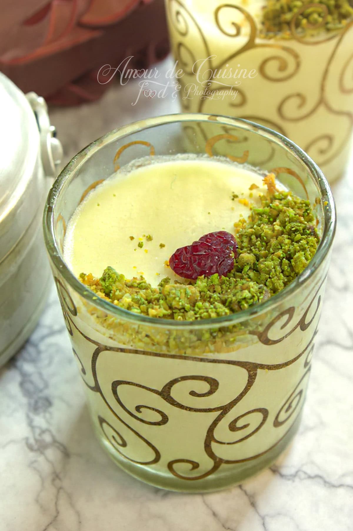 Verrine de mouhalabieh (mahalabiya) bien froide, crème au lait lisse garnie de pistaches concassées et d’une cranberry, sur un plan de travail marbré.