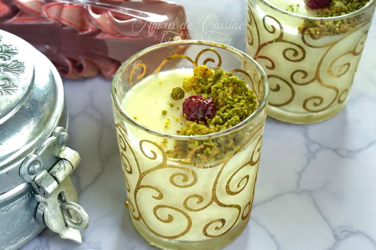 Deux verrines de mouhalabieh (mahalabiya) bien froide, crème au lait garnie de pistaches concassées et d’une cranberry, posées sur un plan de travail marbré avec accessoires en arrière-plan.