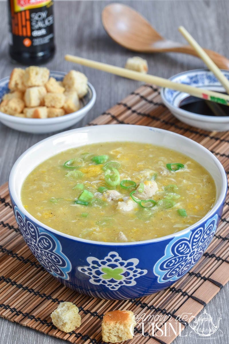 Bowl of Chinese chicken and sweetcorn soup garnished with chopped green onions, served in a decorative blue bowl on a bamboo mat, with soy sauce, chopsticks, croutons, and a wooden spoon in the background.