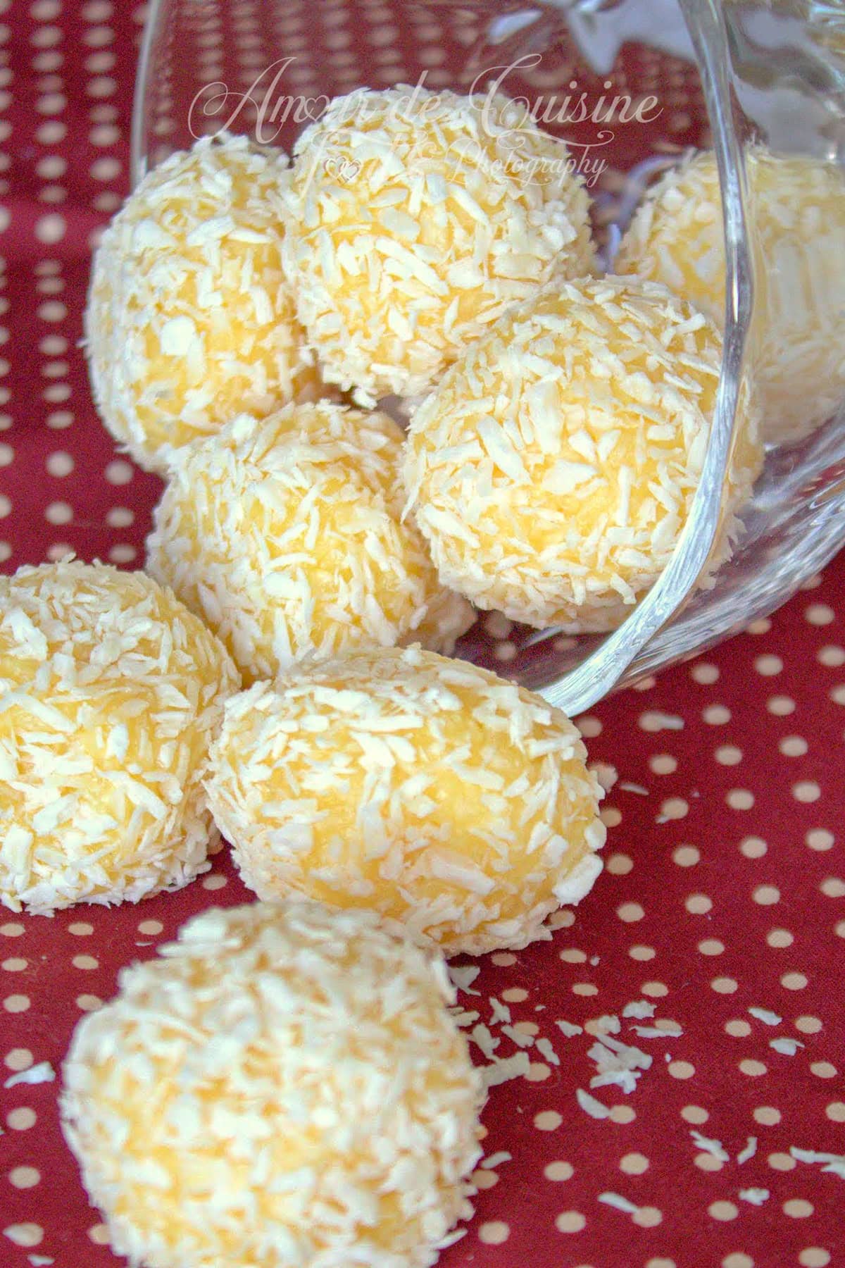 Truffes à la noix de coco enrobées de coco râpée, renversées d’un bol en verre sur un tissu rouge à pois, avec quelques copeaux de coco éparpillés.