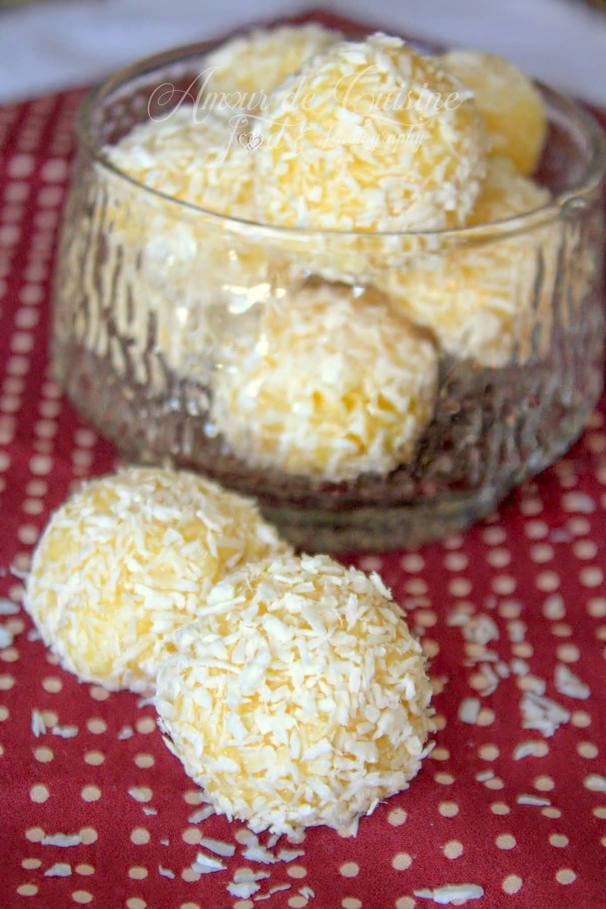 Bol en verre rempli de truffes à la noix de coco roulées dans la coco râpée, avec deux truffes au premier plan sur un tissu rouge à pois et quelques copeaux de coco éparpillés.