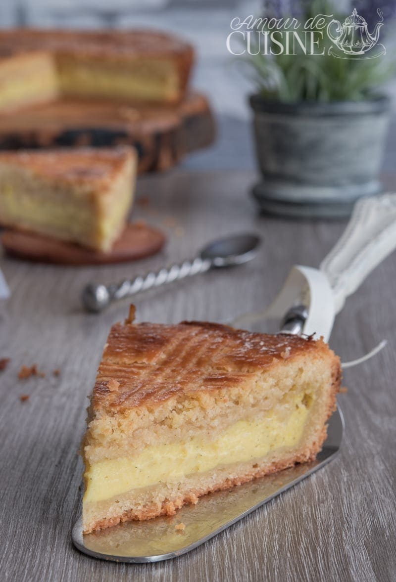Part de gâteau basque à la crème pâtissière posée sur une pelle à gâteau, avec le gâteau entier flou en arrière-plan sur une table en bois.