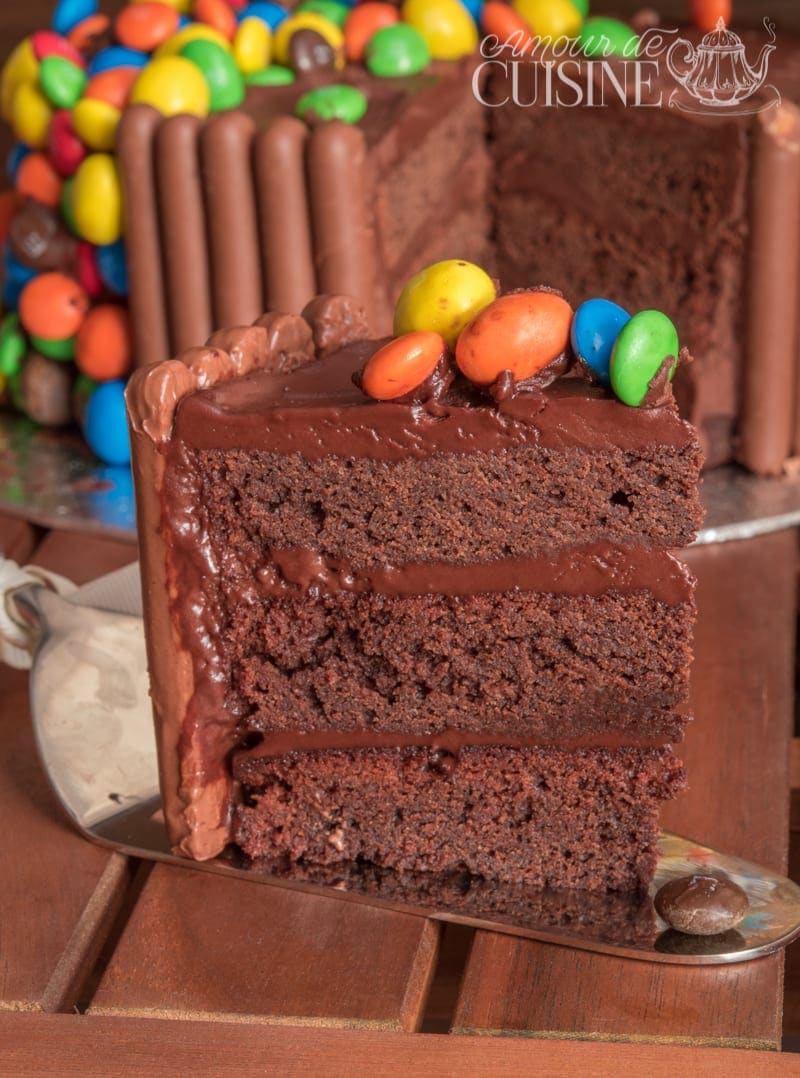Part de gravity cake au chocolat avec trois couches moelleuses et ganache, décorée de M&M’s, avec le gâteau recouvert de Fingers en arrière-plan.