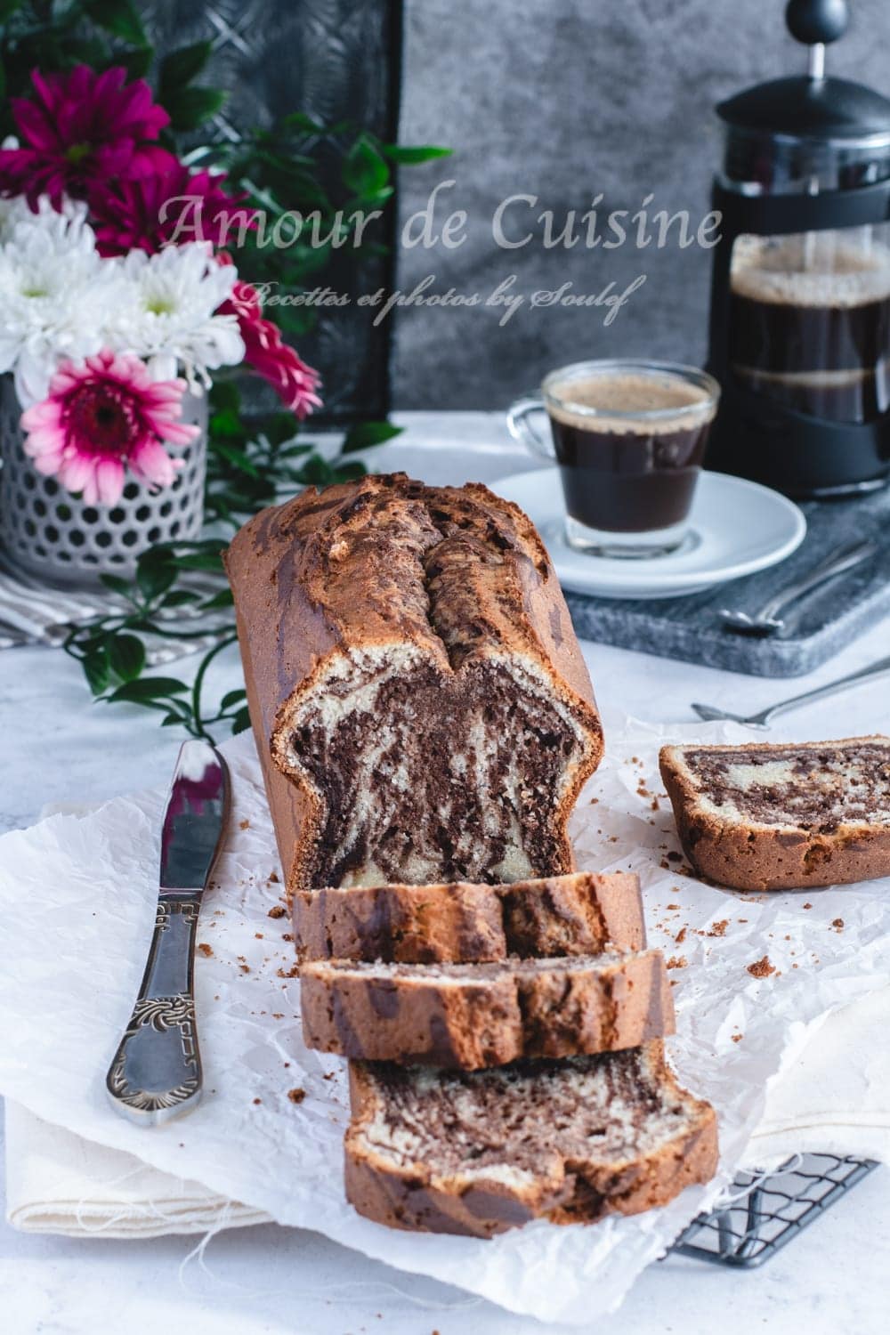Partly sliced chocolate-vanilla marble loaf cake on baking paper with a knife, a cup of coffee and a French press, with flowers in the background.