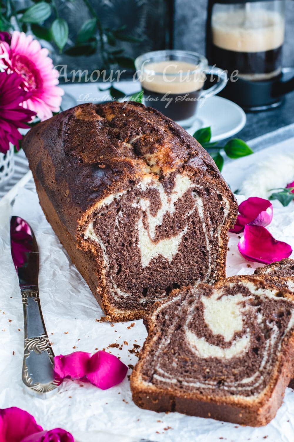 Moist chocolate-vanilla marble loaf cake on baking paper, sliced with one piece in front, two cups of coffee in the background, and scattered flower petals.