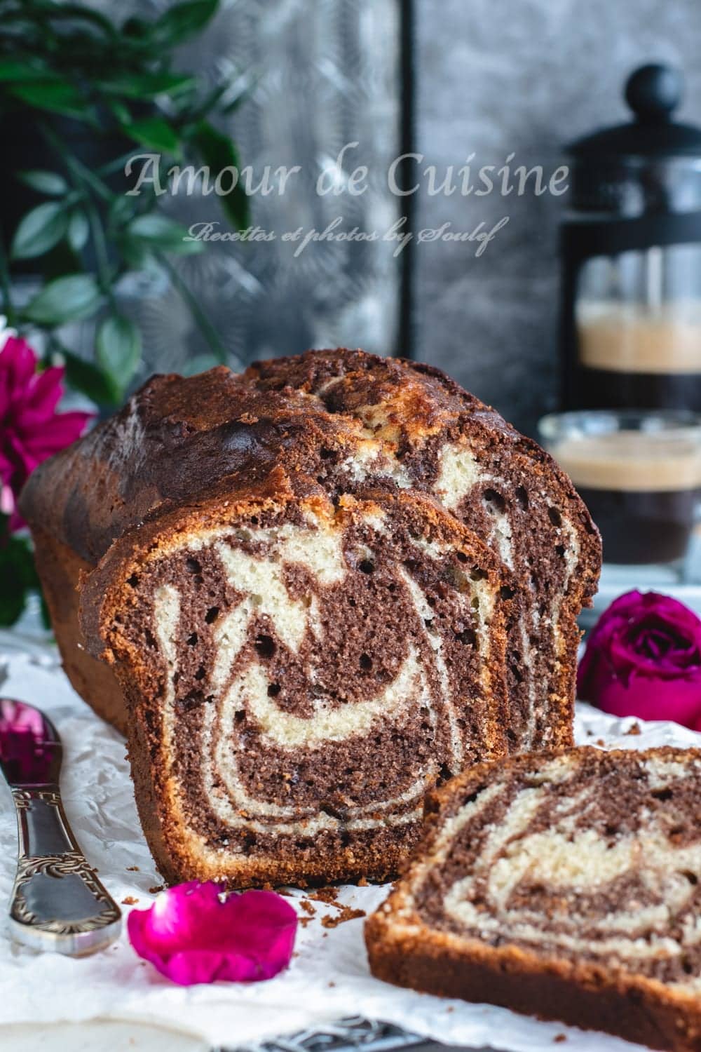 Close-up of a chocolate-vanilla marble loaf cake with one slice in front, rose petals on the table, and two cups of coffee in the background.