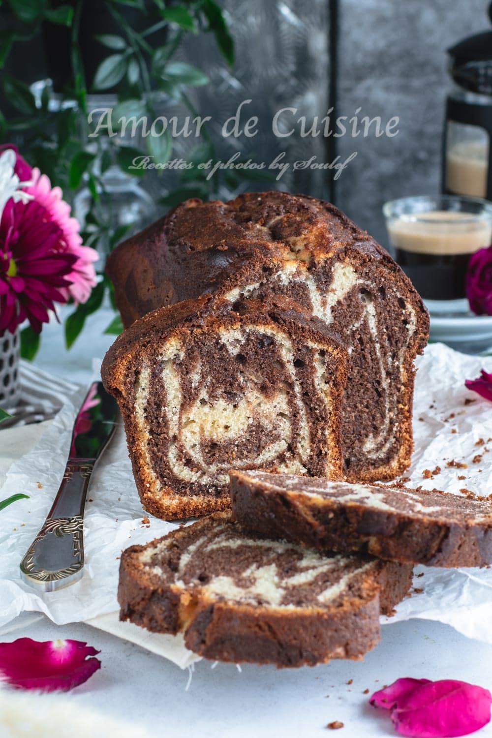 Chocolate-vanilla marble loaf cake sliced on baking paper, with slices in the foreground, pink flowers, and a cup of coffee in the background.