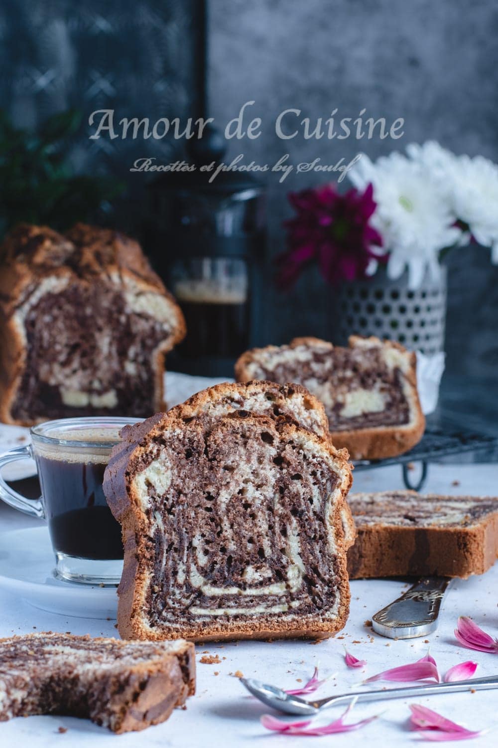 Thick slice of chocolate-vanilla marble loaf cake in front of a cup of coffee, with more slices in the background, pink petals, and a flower bouquet against a grey backdrop.