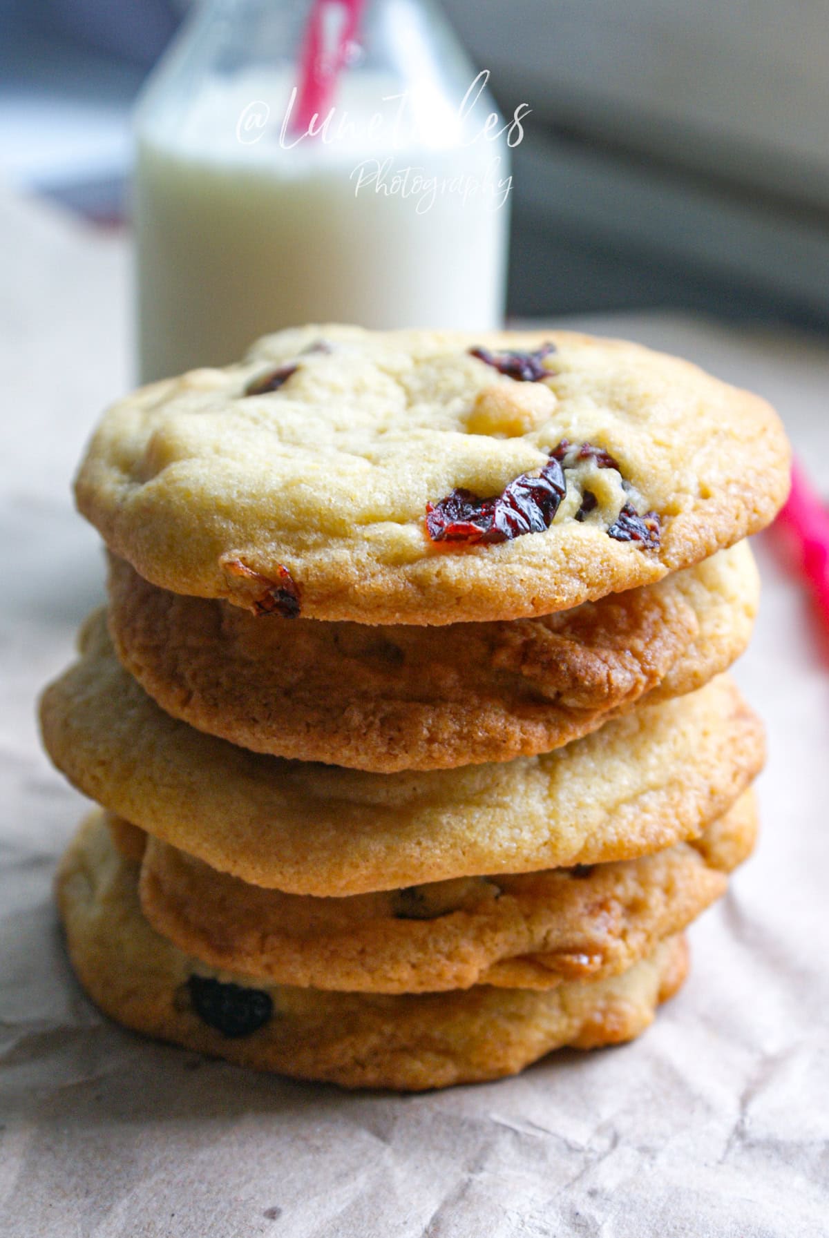 Pile de cookies dorés à la pâte d’amande et cranberries séchées, posée sur du papier cuisson, avec une bouteille de lait en arrière-plan