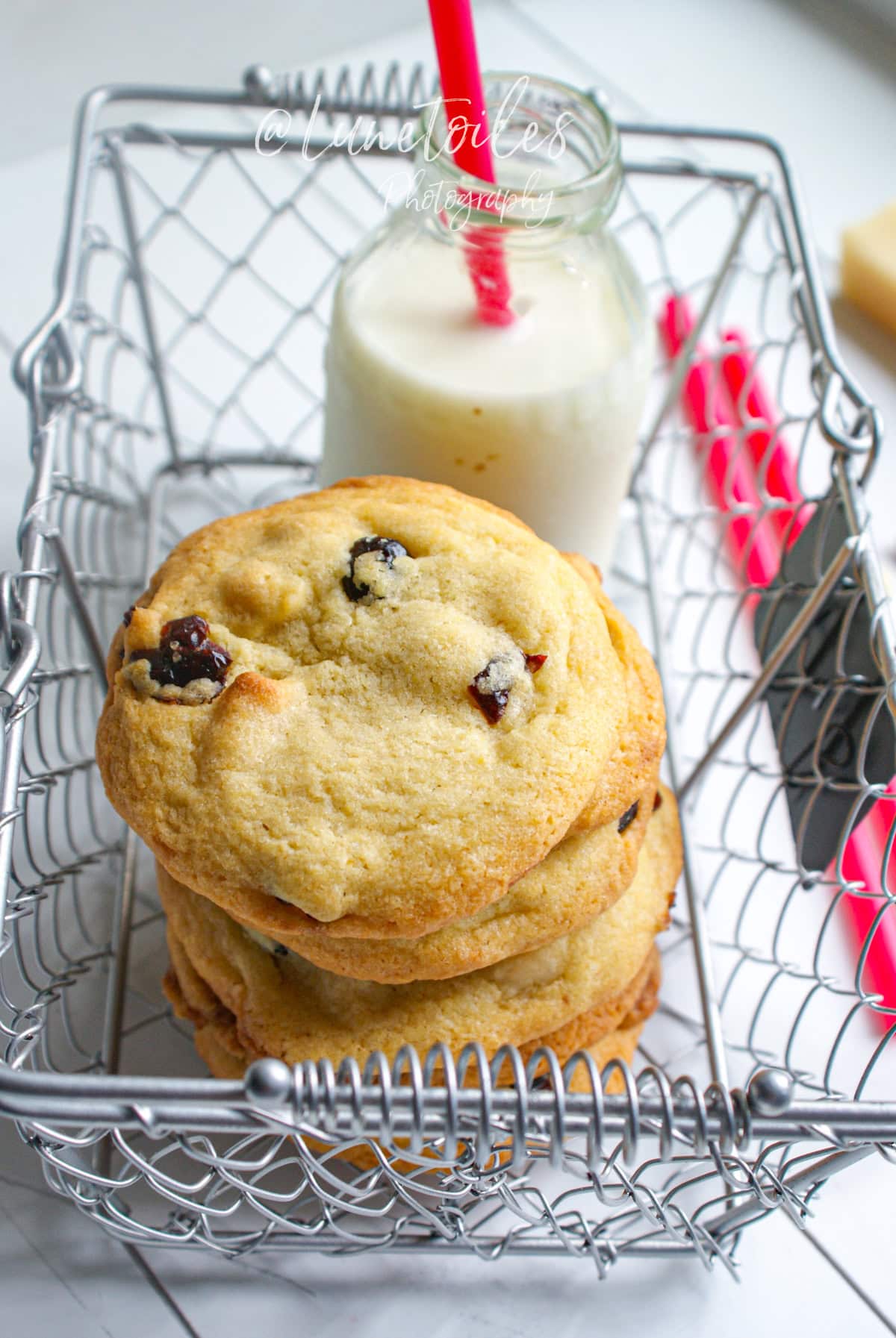 Cookies moelleux à la pâte d’amande et cranberries dans un panier en métal, accompagnés d’une bouteille de lait avec paille rose