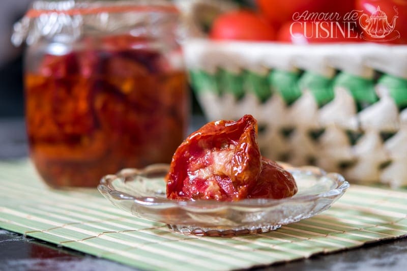 Tender oven-dried tomatoes in olive oil on a glass plate, with a jar of preserves and a soft-focus basket of fresh tomatoes in the background