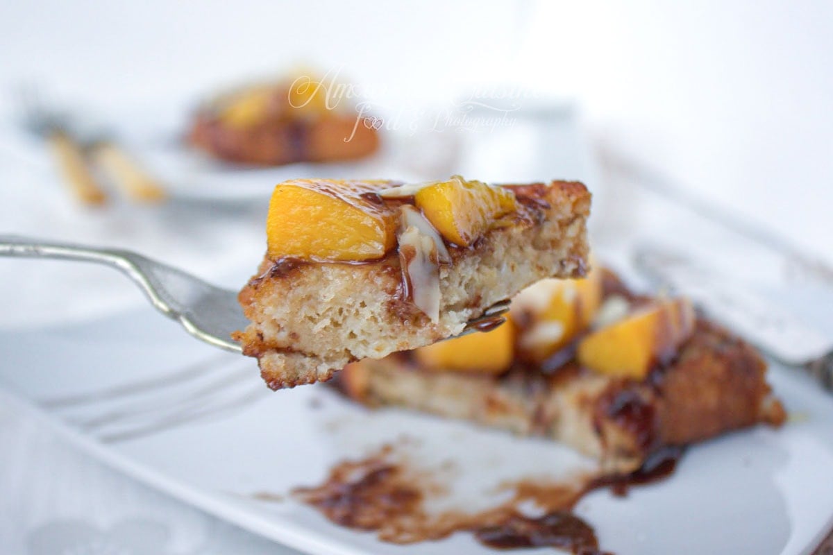 Morceau de brioche perdue moelleuse sur une fourchette, garni de pêches caramélisées, d’amandes effilées et de sauce au chocolat.