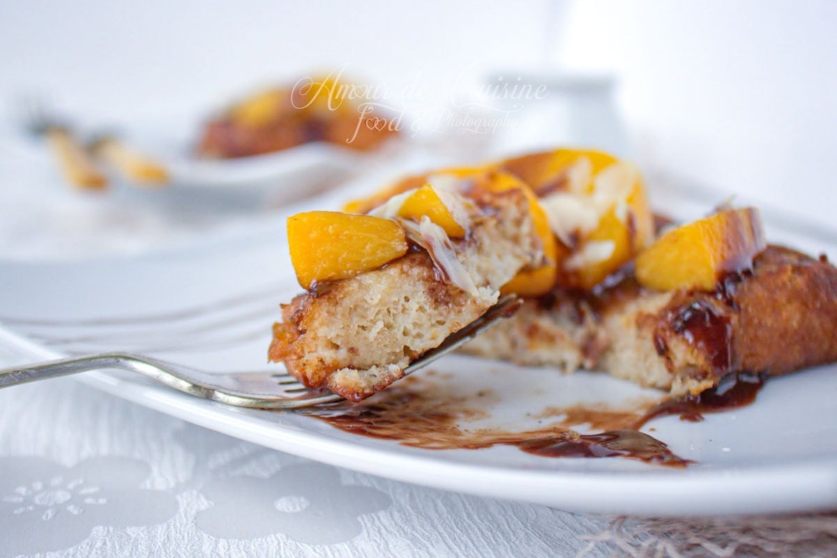 Bouchée de brioche perdue moelleuse aux pêches caramélisées sur une fourchette, avec sauce au chocolat et amandes effilées.