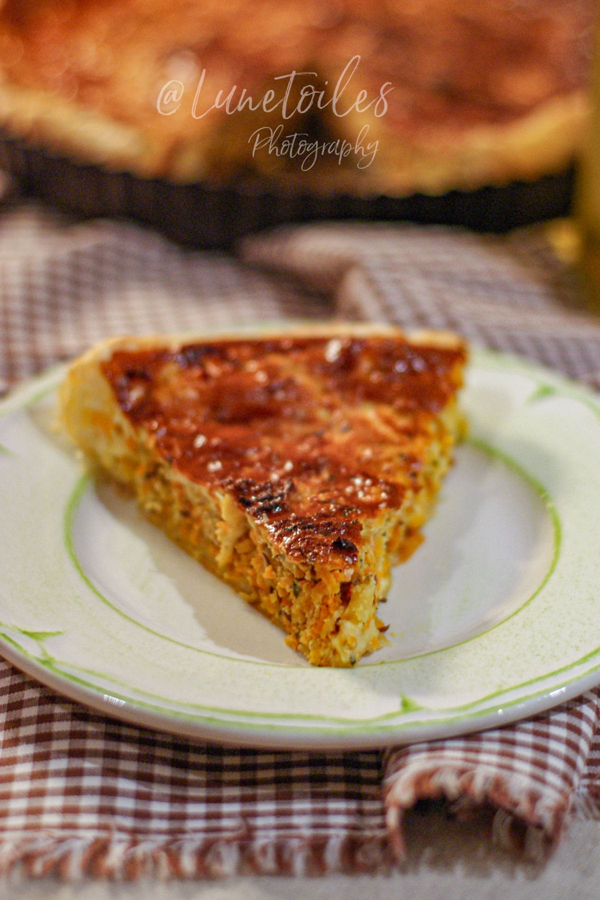 Part de quiche aux carottes au gouda et cumin sur une assiette, avec la quiche entière floutée en arrière-plan.