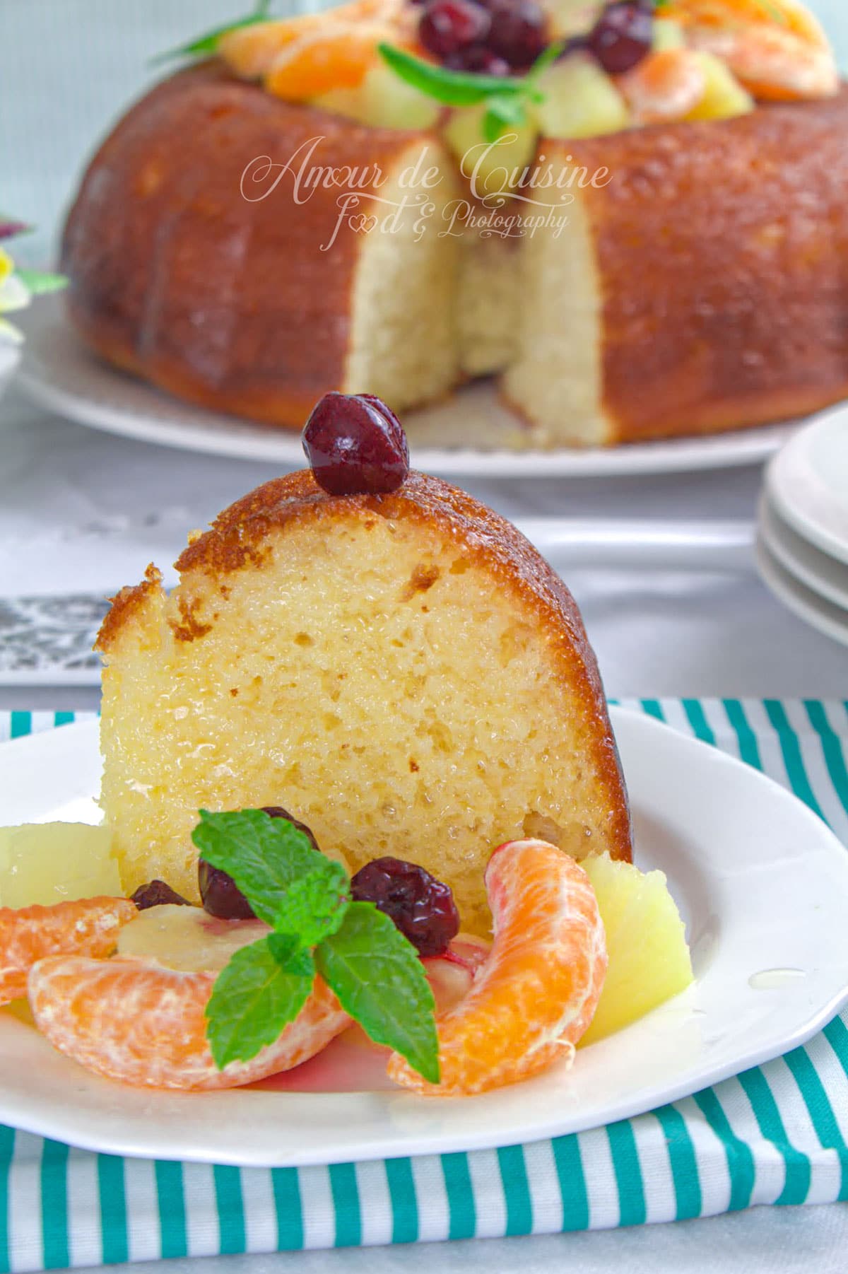 Part de savarin à l’orange sans rhum, bien imbibé et moelleux, servie avec quartiers de mandarine, ananas, cerises et feuilles de menthe, avec le gâteau entier en arrière-plan.