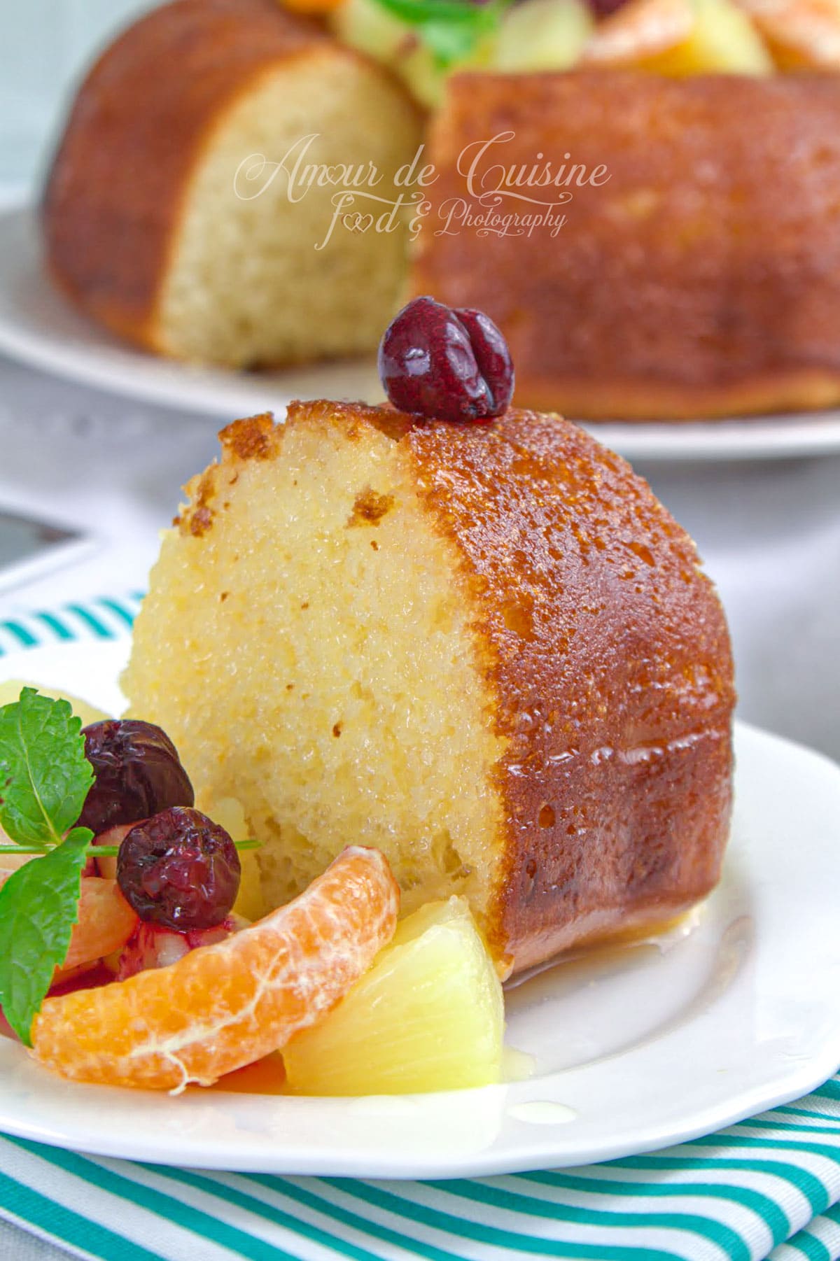 Part de savarin à l’orange sans rhum, moelleux et bien imbibé, servie avec mandarine, ananas, cerises et menthe, avec le gâteau entier en arrière-plan.