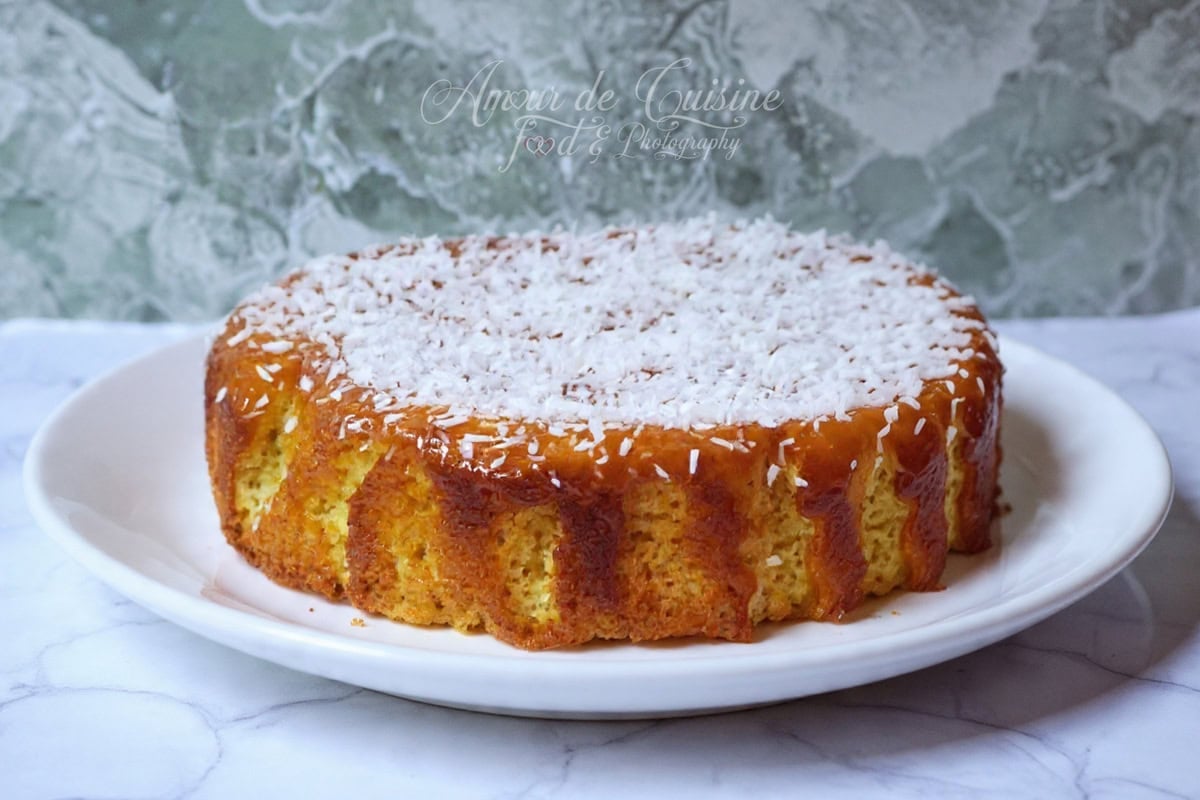 Gâteau à la noix de coco entier, nappé de lait concentré sucré qui coule sur les bords, puis saupoudré de noix de coco râpée, présenté sur une assiette blanche posée sur un plan de travail effet marbre.