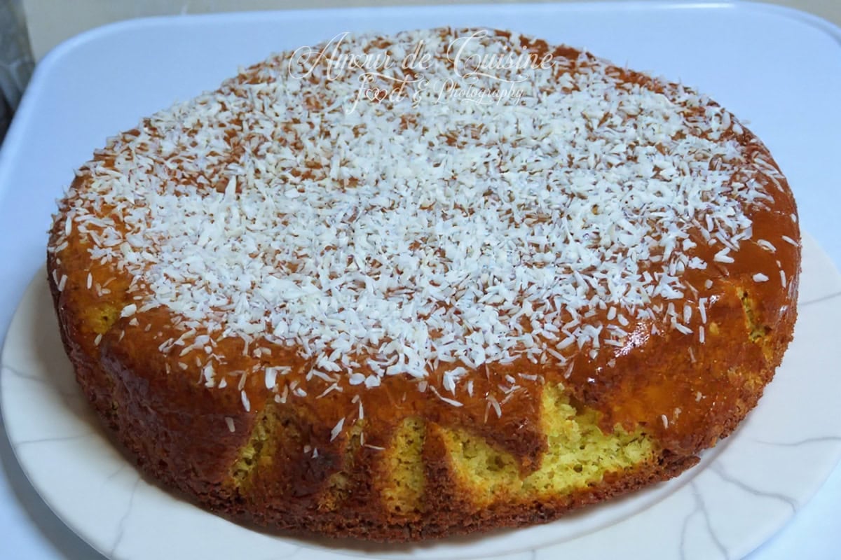 Gâteau à la noix de coco entier, doré et brillant, nappé puis généreusement saupoudré de noix de coco râpée, présenté sur une assiette blanche.