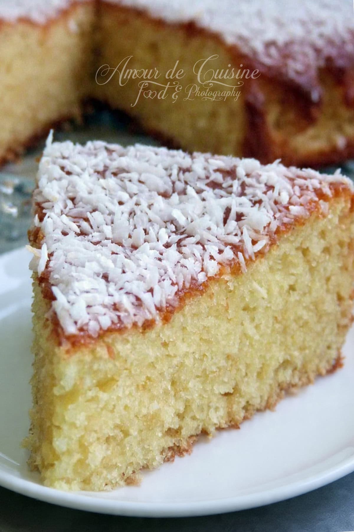 Part de gâteau moelleux à la noix de coco, recouverte de noix de coco râpée, servie sur une assiette blanche, avec le reste du gâteau flouté en arrière-plan.