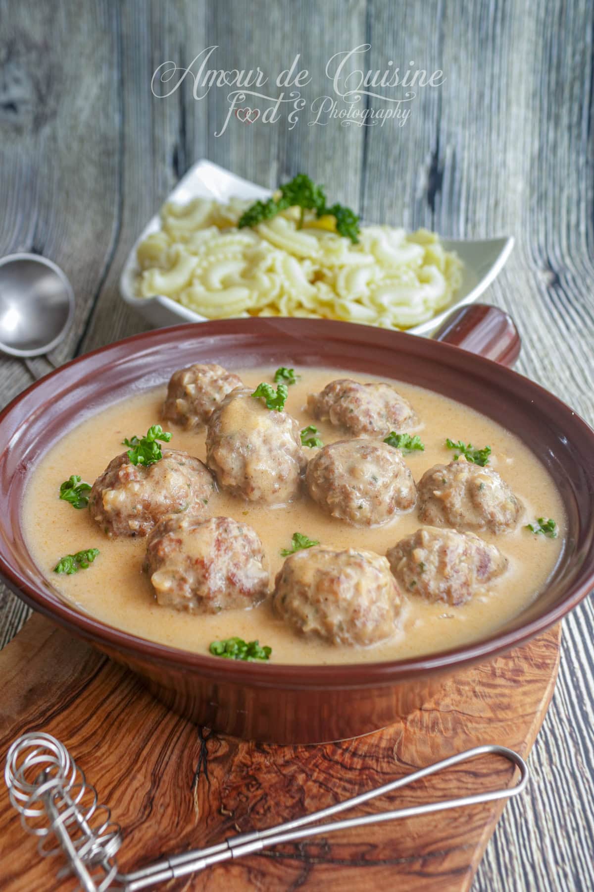 Dish of Swedish meatballs served in a creamy sauce, garnished with fresh parsley, with pasta in the background.
