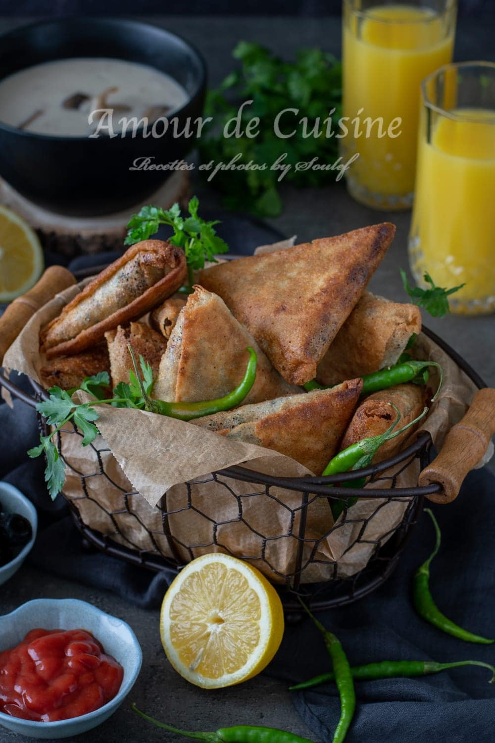 Panier de bricks (boureks) dorés et croustillants, en triangles et roulés, servis avec piments verts, persil, citron, olives, sauce tomate, un bol de soupe et des verres de jus d’orange en arrière-plan.