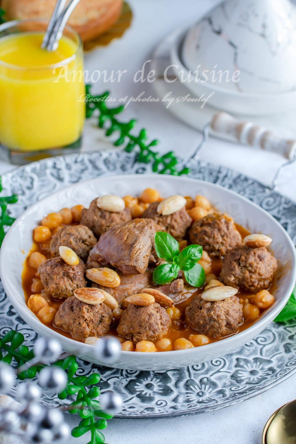 Bowl of Algerian mtewem in red sauce with meatballs, lamb chunks, chickpeas, almonds, and basil, served on a patterned plate with a tagine dish in the background.