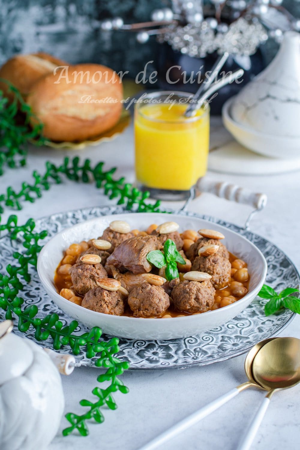 Plate of Algerian mtewem in red sauce with meatballs, chickpeas, toasted almonds, and basil, set on a patterned serving tray, with matlouh bread, a tagine dish, and a drink in the background.