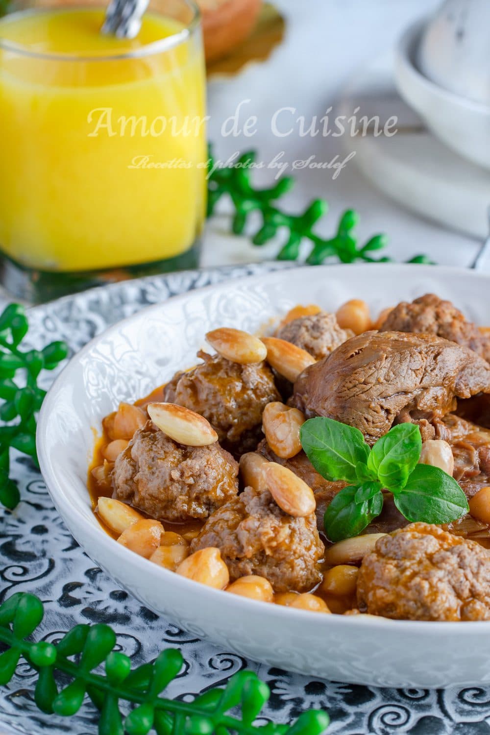 Close-up of a bowl of Algerian mtewem in red sauce with meatballs, lamb pieces, chickpeas, toasted almonds, and basil, served on a patterned plate with a glass of yellow drink in the background.