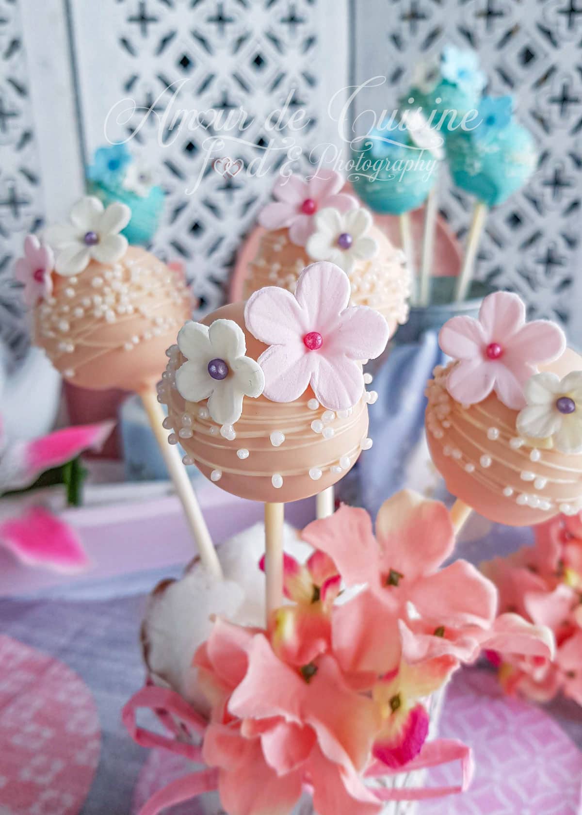 Pastel pink cake pops decorated with tiny sugar flowers and white pearls, displayed on sticks with a delicate floral background, creating a soft and festive atmosphere.