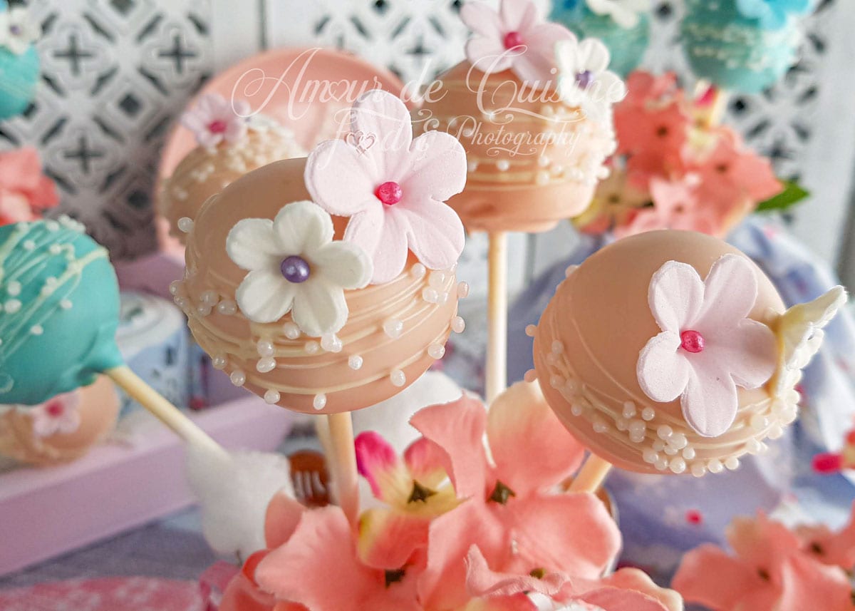 Pink cake pops decorated with sugar flowers and small white pearls, displayed on sticks among pink blossoms, with other colourful cake pops in the background in a soft, festive setting.