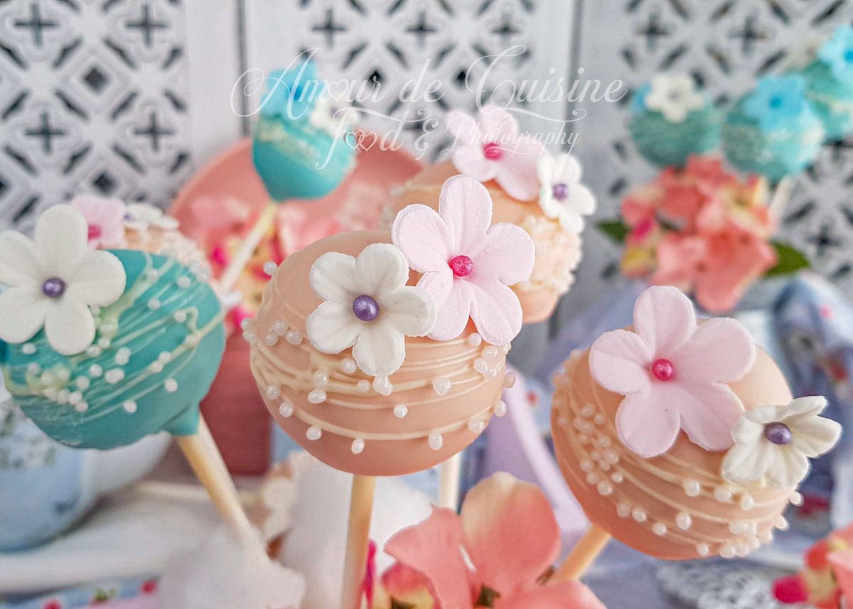 Pastel cake pops decorated with sugar flowers and white pearls, displayed on sticks among pink blossoms, with colourful cake pops blurred in the background for a soft, festive presentation.