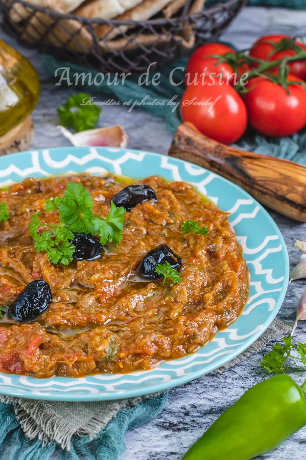Assiette de zaalouk marocain aux aubergines, décoré d’olives noires et de coriandre, présenté avec des tomates fraîches, ail et pain maison en arrière-plan