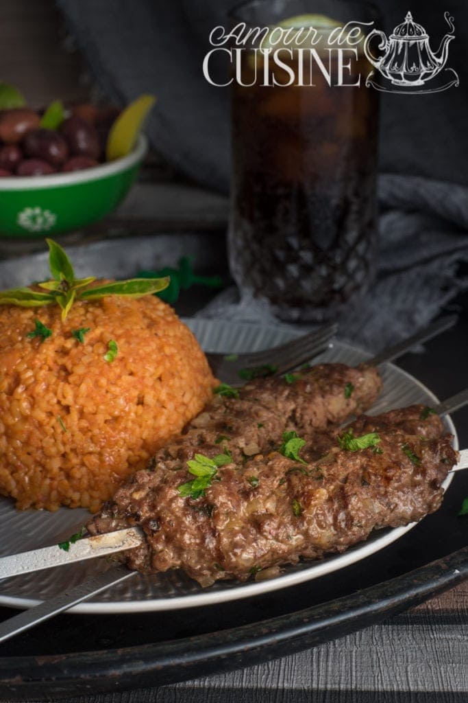 Grilled kefta skewers served with tomato bulgur and garnished with fresh parsley, next to a glass of iced drink and a bowl of olives